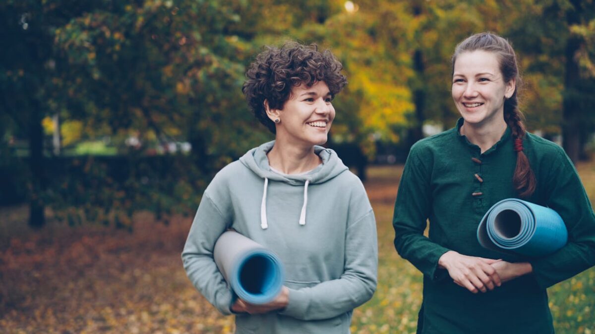 Two women holding yoga mats, smiling and walking in a park during autumn.