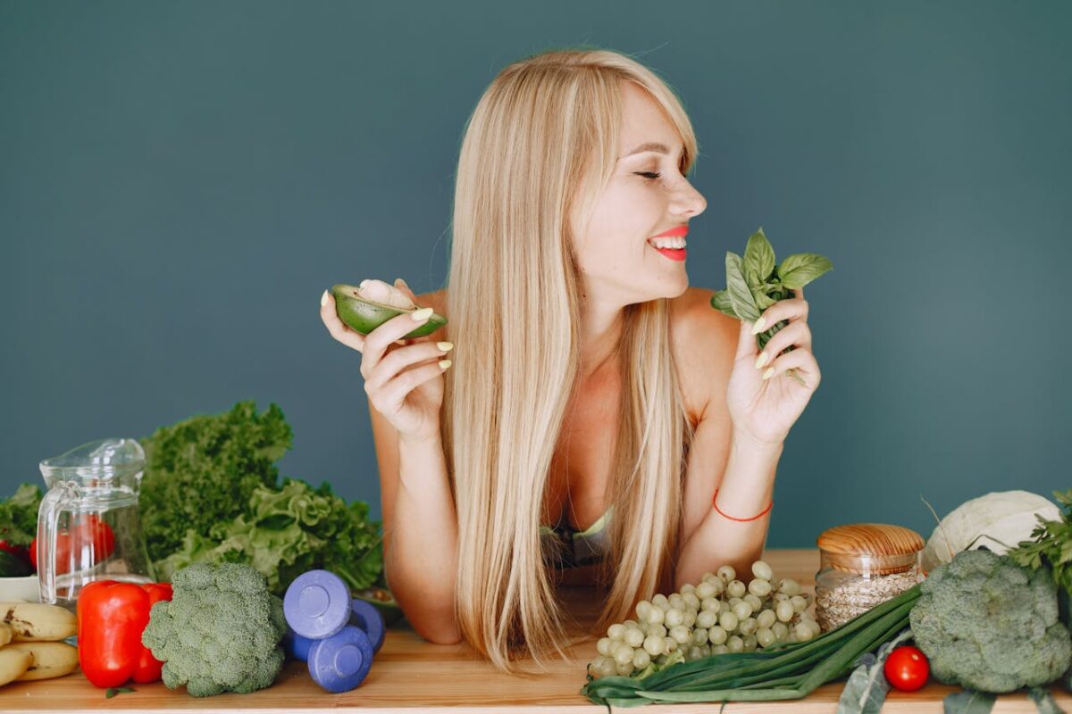 A smiling woman holding avocado and basil, surrounded by fresh vegetables and fruits for healthy eating.