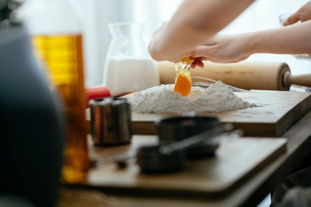 Unrecognizable cook breaking egg into pile of flour on wooden board while preparing dough at table in kitchen against blurred background