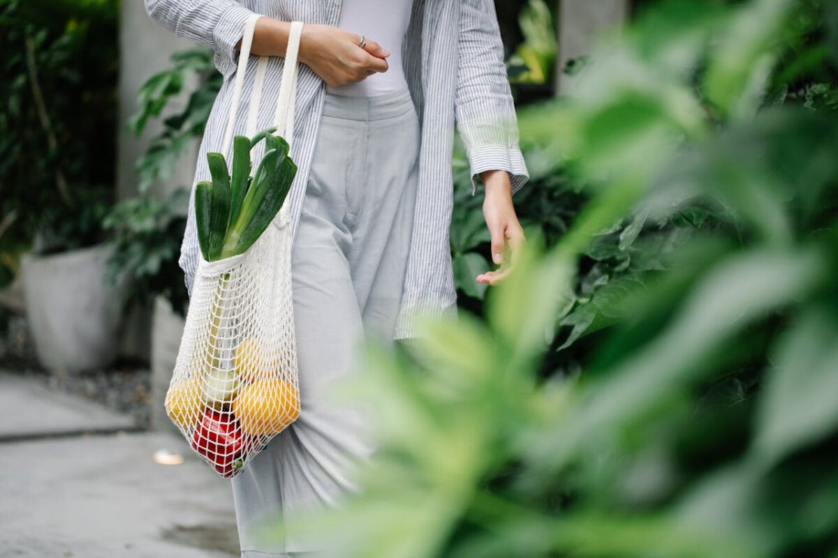 Crop anonymous female wearing casual outfit carrying net grocery bag with ripe vegetables including leek and capsicums in garden