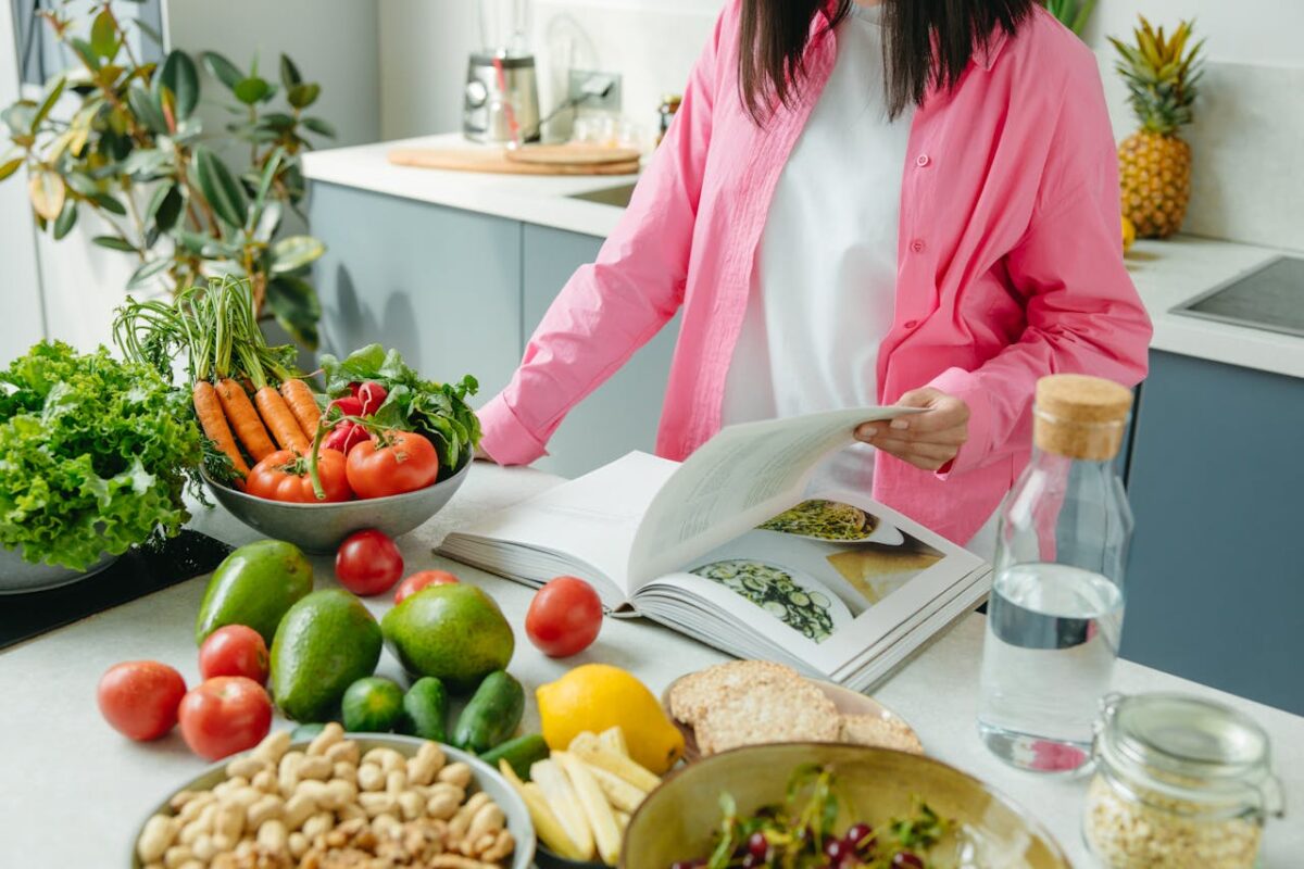 A person browsing a cookbook surrounded by fresh vegetables in a bright kitchen setting.